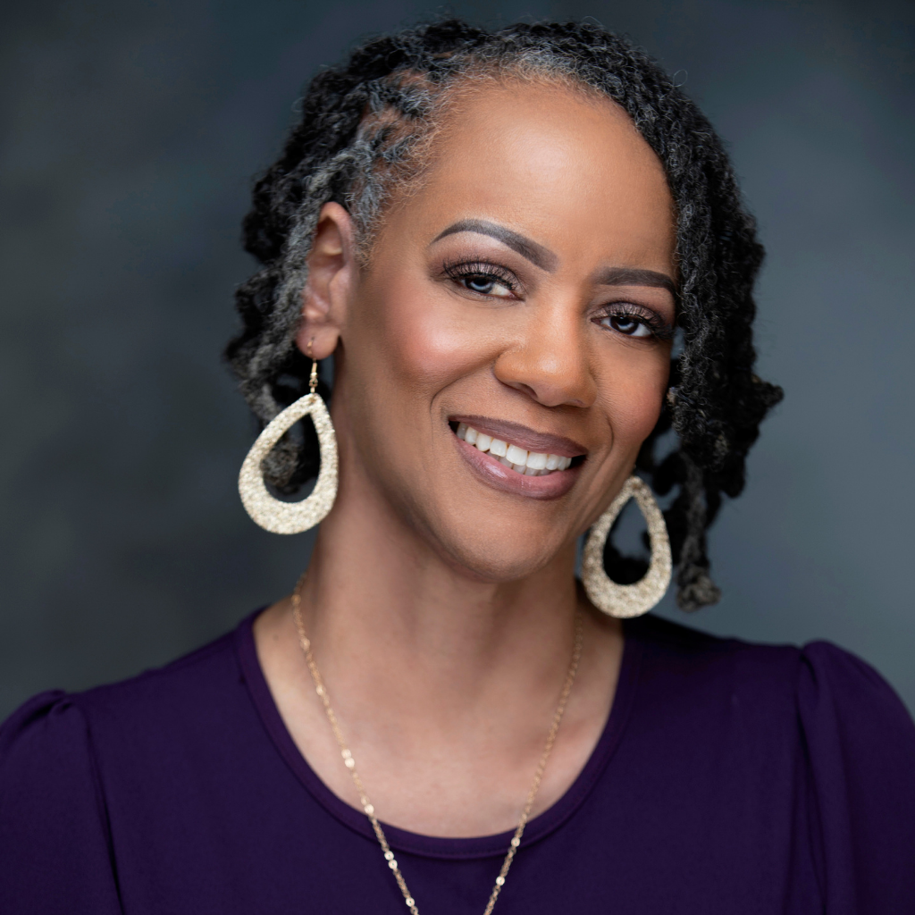 Portrait of a smiling woman with textured short dreadlocks featuring gray strands, wearing large glittery teardrop earrings and a purple top.”] ,