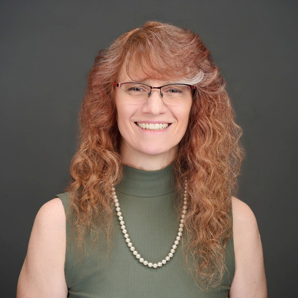 Smiling woman with curly red hair and glasses, wearing a green sleeveless top and a pearl necklace against a gray background.