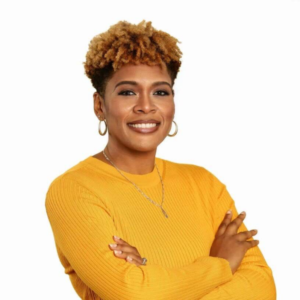 Smiling woman with short curly blond hair in a mustard yellow ribbed sweater, arms crossed and wearing hoop earrings.
