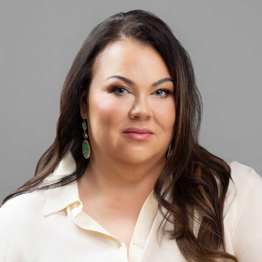 Head-and-shoulders portrait of a smiling woman with long dark hair, wearing a cream blouse and green earrings against a gray backdrop.