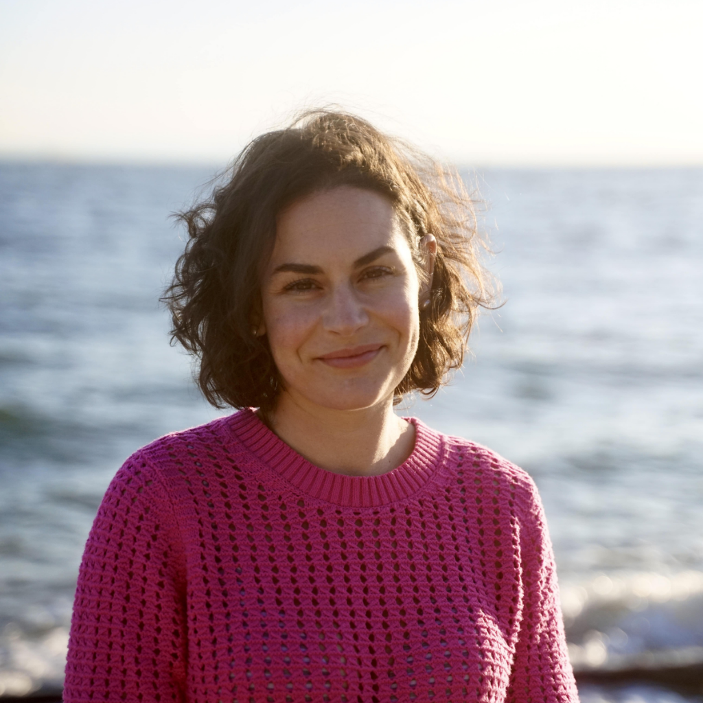Woman with short curly brown hair smiling at the camera on a beach, wearing a pink crochet-knit sweater.