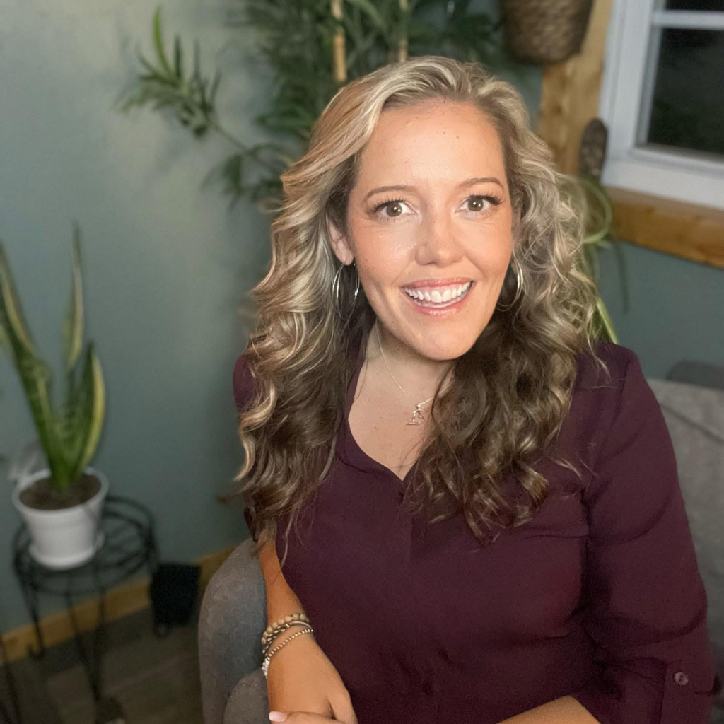 Smiling woman with wavy blonde hair in a maroon blouse sits indoors, plants in the background.