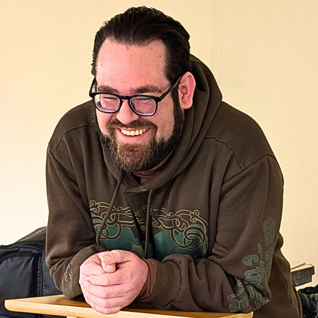 Smiling man with dark hair and beard wearing glasses, in a brown hoodie, leaning on a wooden table against a beige wall.