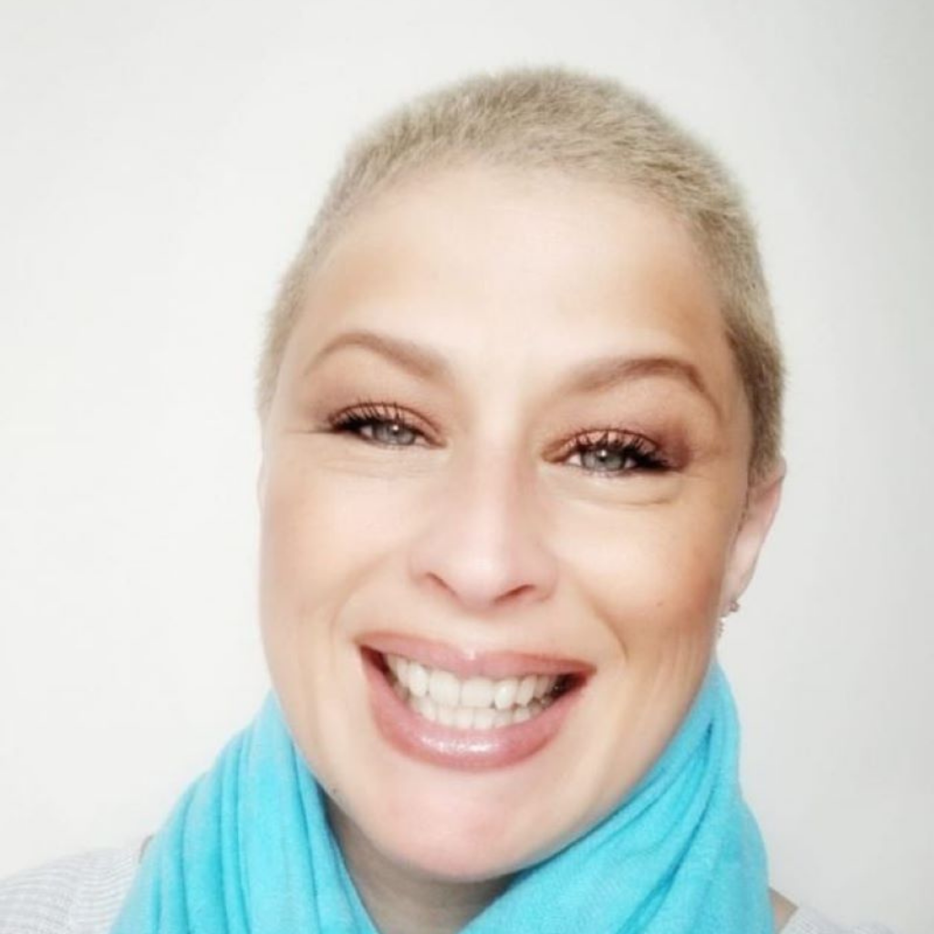 Smiling portrait of a woman with short blond hair wearing a turquoise scarf against a light background.
