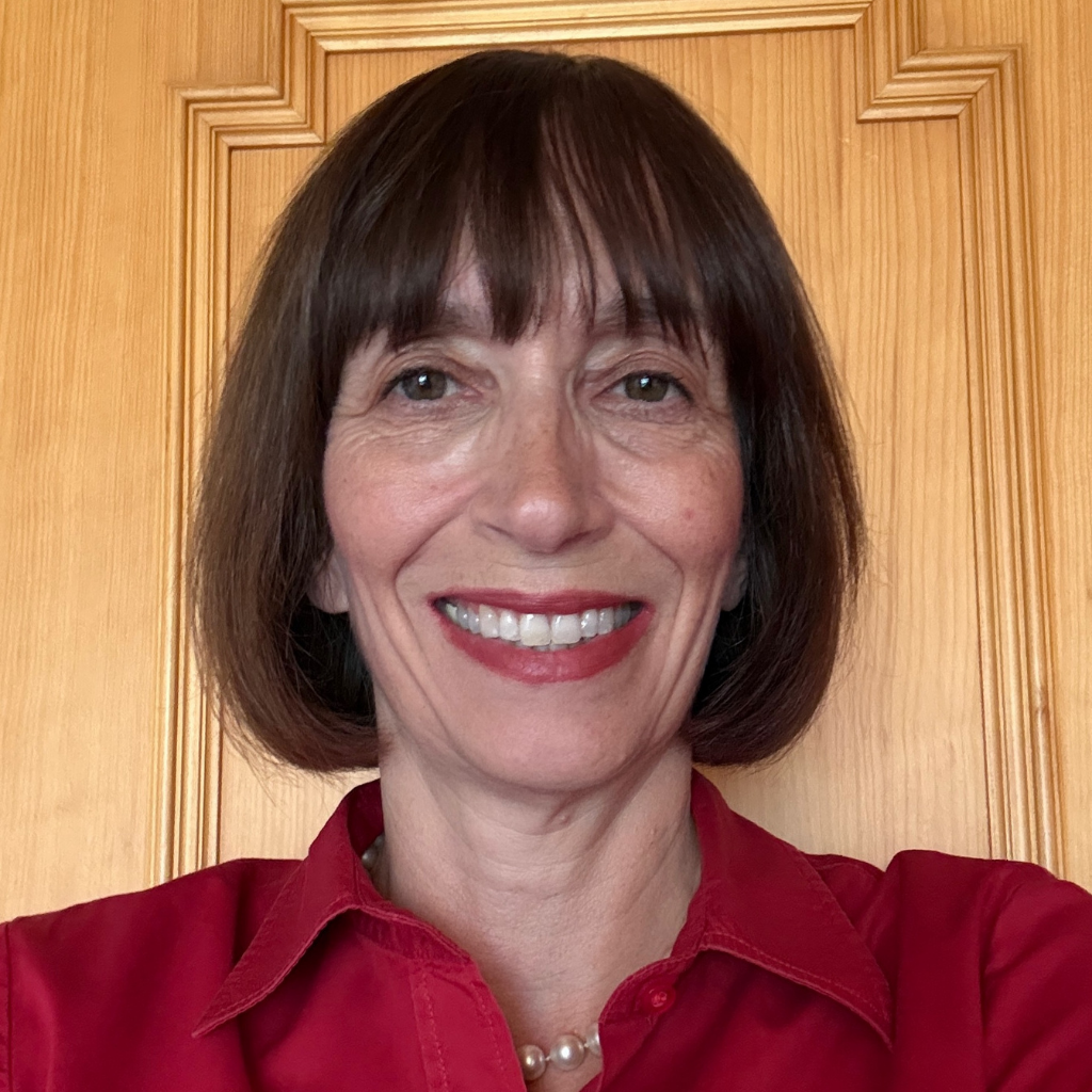 Smiling woman with short dark hair wearing a red blouse and pearl necklace, standing in front of a wooden door