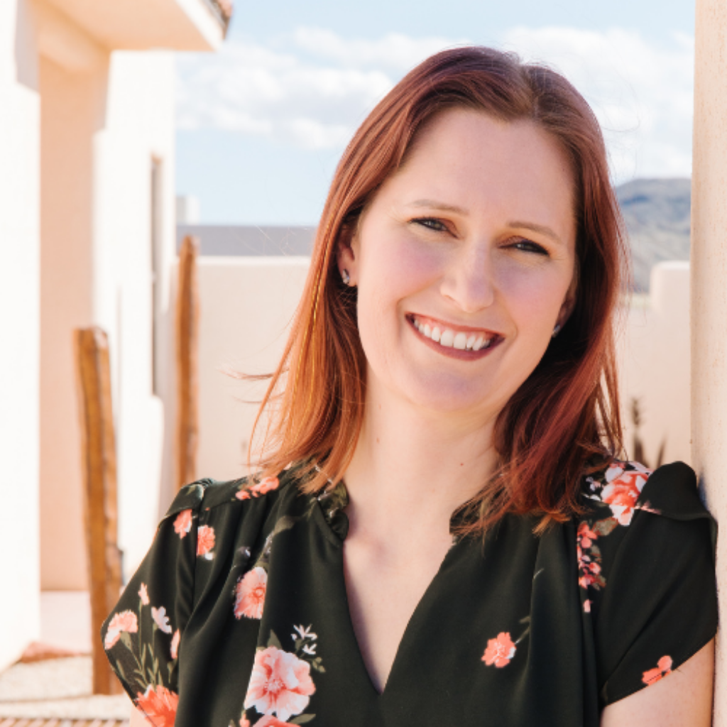 Smiling woman with red hair leaning against a light wall outdoors, wearing a dark floral dress.