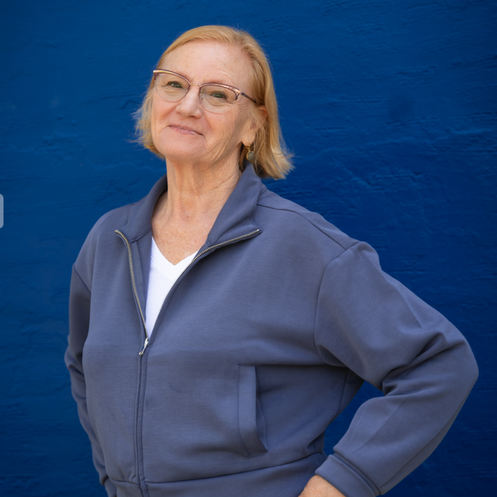 Smiling older woman with short blond hair and glasses, wearing a blue zip-up jacket against a blue wall.