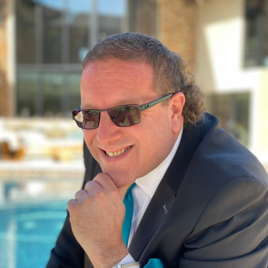 Smiling man in a black suit and teal tie, wearing sunglasses, resting his chin on his hand by a poolside.