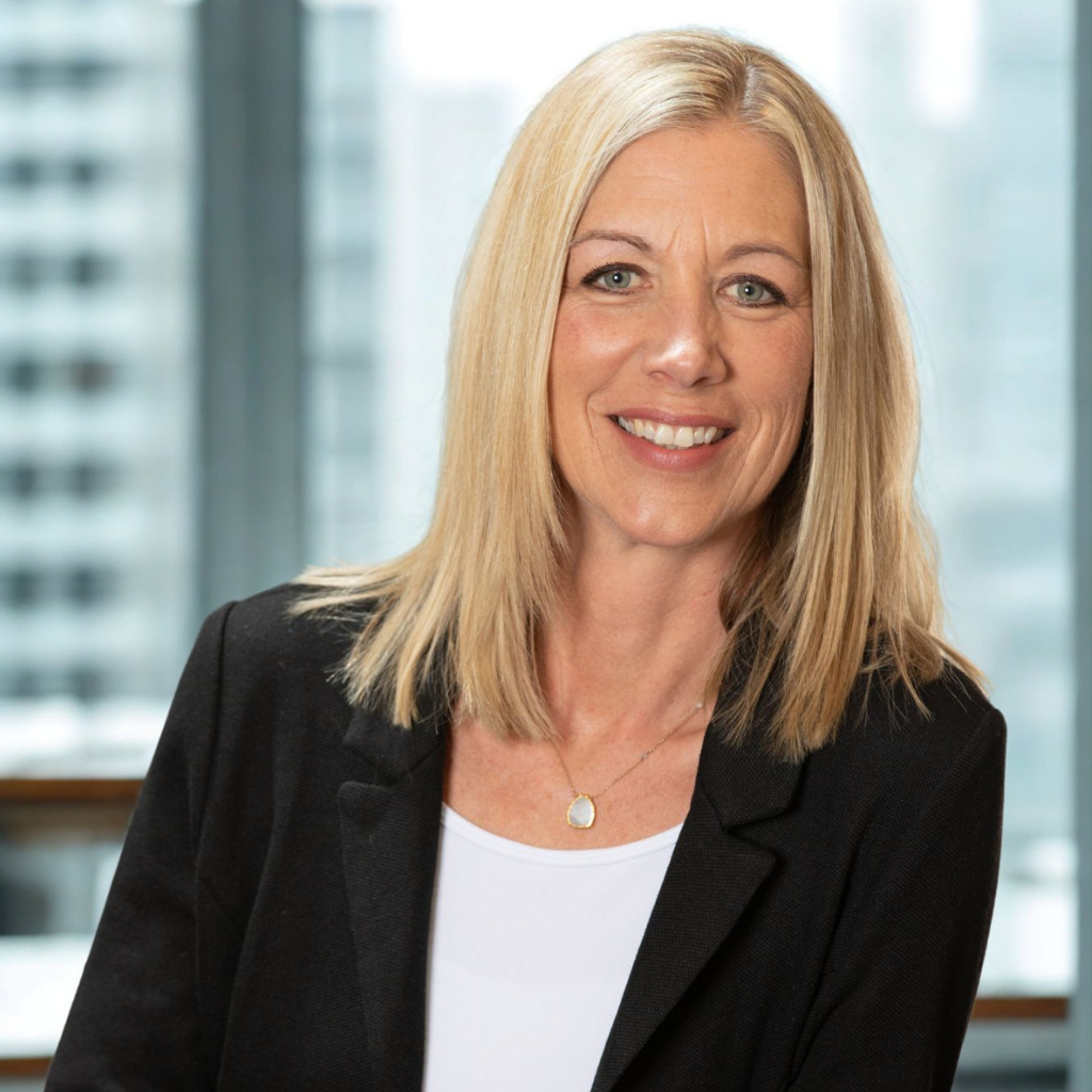 Professional headshot of a smiling blonde woman in a black blazer and white top, wearing a gold pendant necklace, in an office setting.