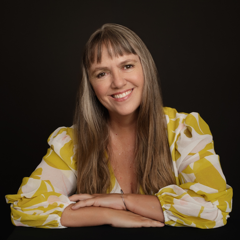 Smiling woman with long light brown hair in a yellow floral blouse sits with arms crossed on a table against a dark background.