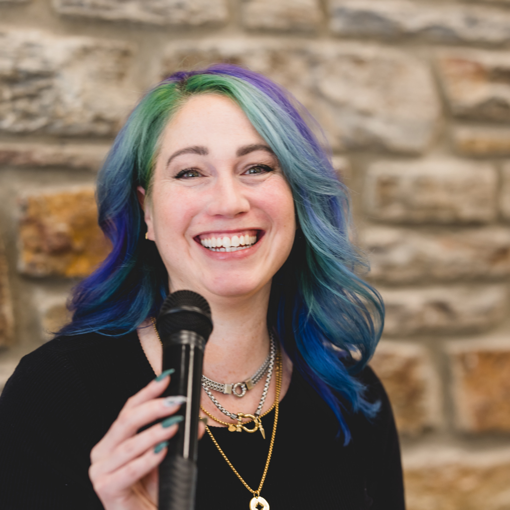 Smiling woman with multicolored hair holding a microphone against a stone wall backdrop.