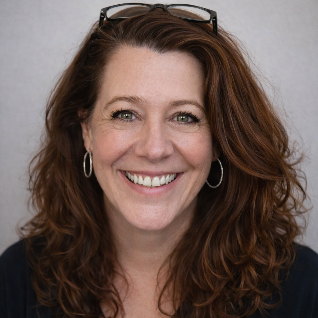 Smiling woman with wavy red hair, glasses on her head, hoop earrings, against a gray backdrop.