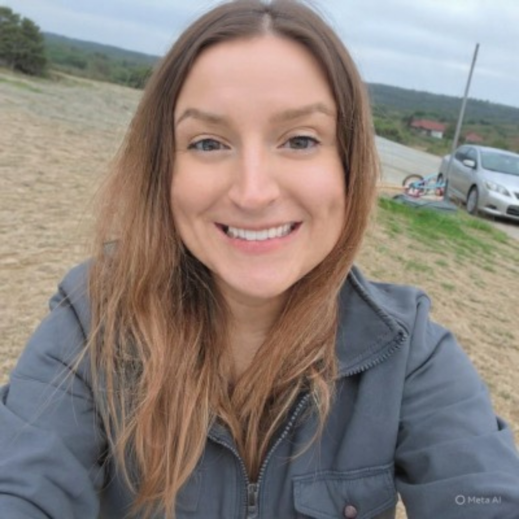 Close-up portrait of a smiling woman outdoors in a gray jacket, with a dirt field and distant hills in the background.