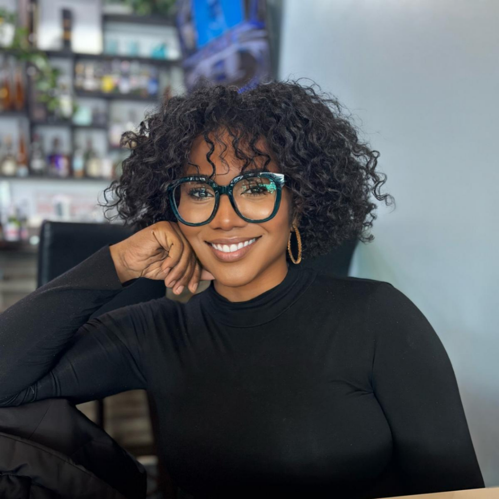 Smiling woman with curly hair and teal glasses rests her chin on her hand in a black turtleneck, seated in a bright indoor space.