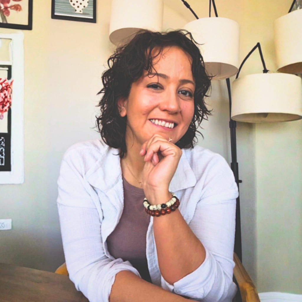 Smiling woman with short curly hair seated indoors, resting her chin on her hand, wearing a light shirt and beaded bracelets.