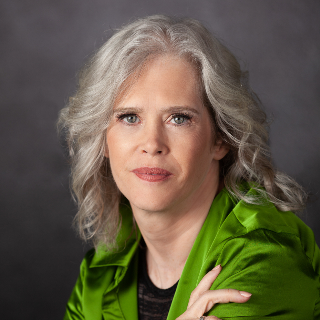 Portrait of a mature woman with silver hair and green satin blouse looking at the camera in a neutral studio backdrop at chest level.